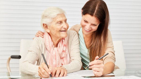 Eine ältere Dame wird durch eine Mitarbeiterin einer Rehaklinik bei dem Ausfüllen eines Sudokus unterstützt.