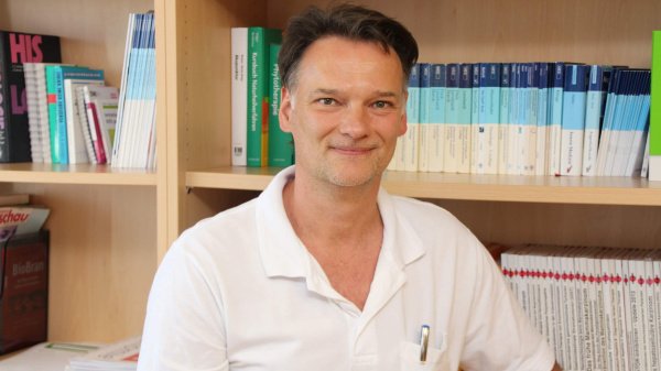 Gastroenterologe im weißen Shirt vor Bücherregal mit weiß-blauen Fachbüchern