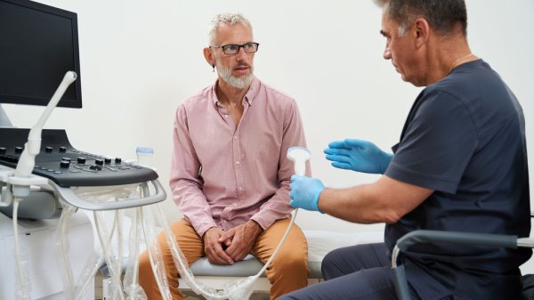 Ein Mann sitzt auf einer Liege im Krankenhaus, während ein Arzt mithilfe eines Ultraschallgeräts die bevorstehende Untersuchung auf ein Aortenaneurysma erklärt.