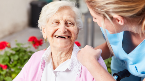 Ältere Patientin lacht Pflegerin an, die sich über den Stuhl herunter beugt