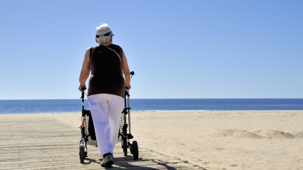 Frau spaziert mit Rollator an einem Strand in der Sonne.