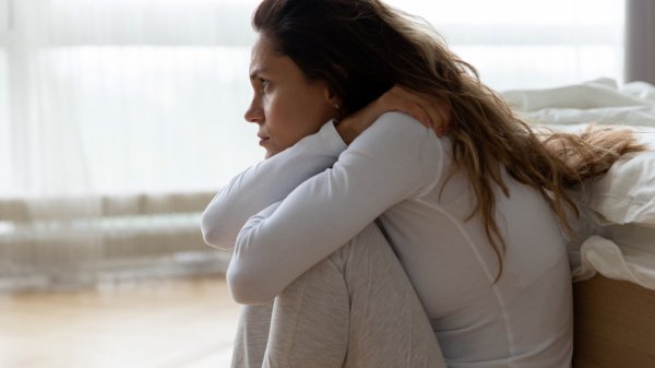 Eine verängstigte Frau sitzt in der Hocke auf dem Boden vor einem Bett, sichtlich belastet von einer Angststörung.