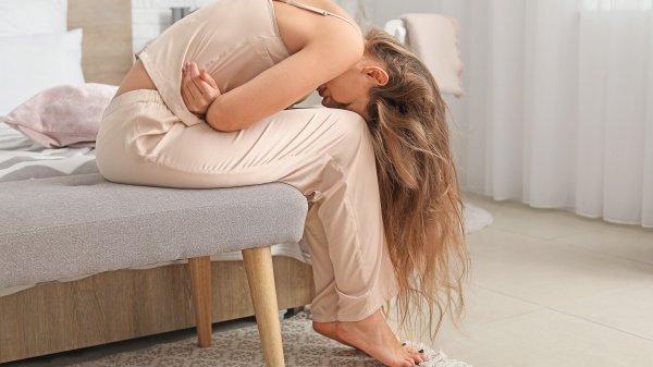 Frau mit Unterleibsschmerzen sitzt gekrümmt auf einen Stuhl.