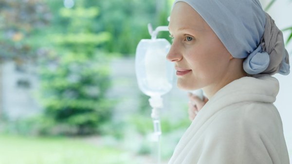 Frau mit Kopfbedeckung berührt einen Infusionsständer mit Blick aus dem Fenster ins Grüne und erholt sich von Chemotherapie.