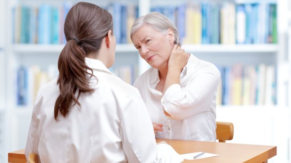 Gespräch zwischen Ärztin und Patientin, die sich den Nacken vor Schmerzen hält.