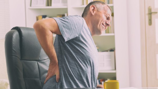 Mann sitzt im Bürostuhl am Arbeitsplatz und hält sich vor Rückenschmerzen den Lendenbereich.