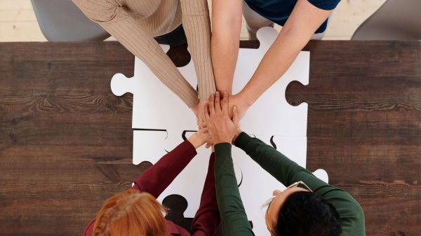 4 Personen halten über ein Puzzle die Hände zusammen. Motto: Hand in Hand arbeiten.