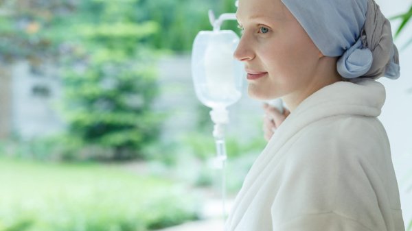 Frau mit Kopfbedeckung berührt einen Infusionsständer mit Blick aus dem Fenster ins Grüne und erholt sich von Chemotherapie.