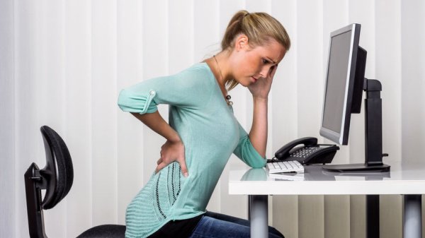 Frau hält sich den Rücken vor Schmerzen und sitzt am Schreibtisch vor einem Computer.