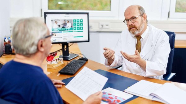 Arzt und Patient im Gespräch gegenüber am Schreibtisch mit medizinischen Unterlagen.
