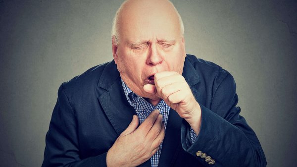 Ein älterer Mann im Anzug hält sich die Brust und eine Faust vor den Mund aufgrund eines Hustenanfalls durch COPD.