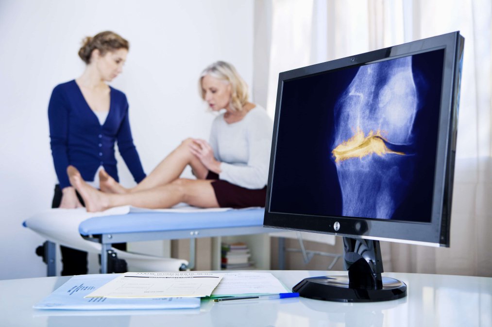 Frau auf Patientenliege mit Blick auf angewinkeltes Knie und Ärztin daneben.