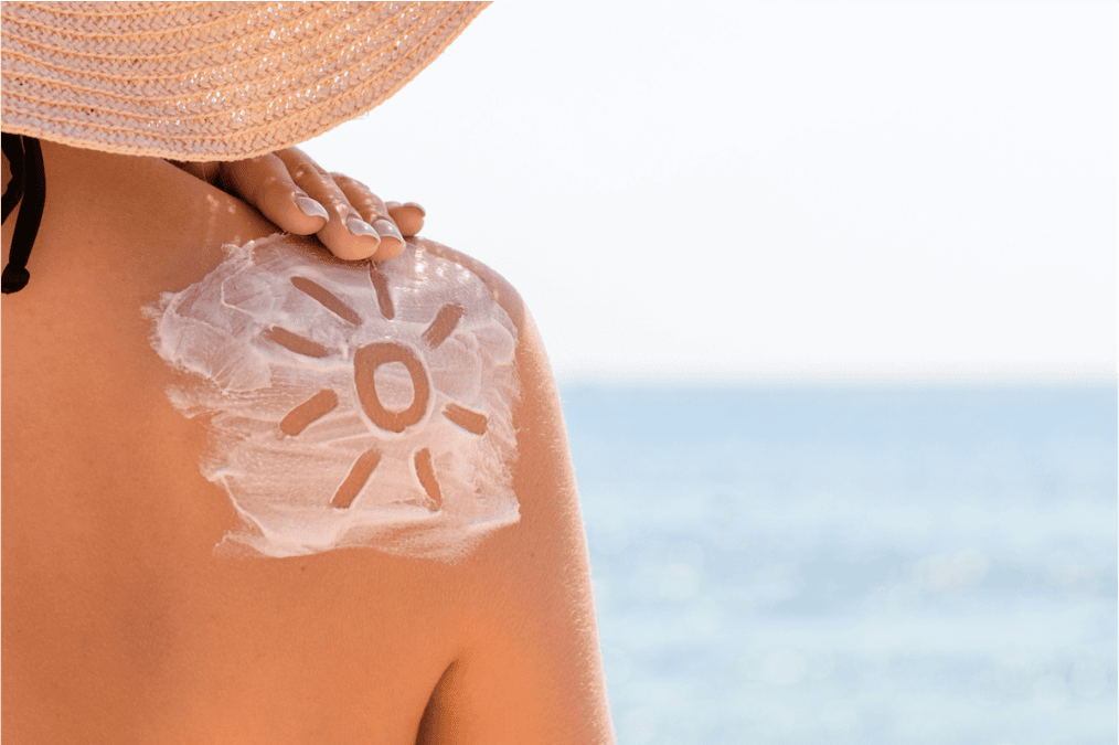 Eine Frau steht am Meer mit Blick auf eine Sonne mit Sonnencreme auf ihrem Schulterblatt.