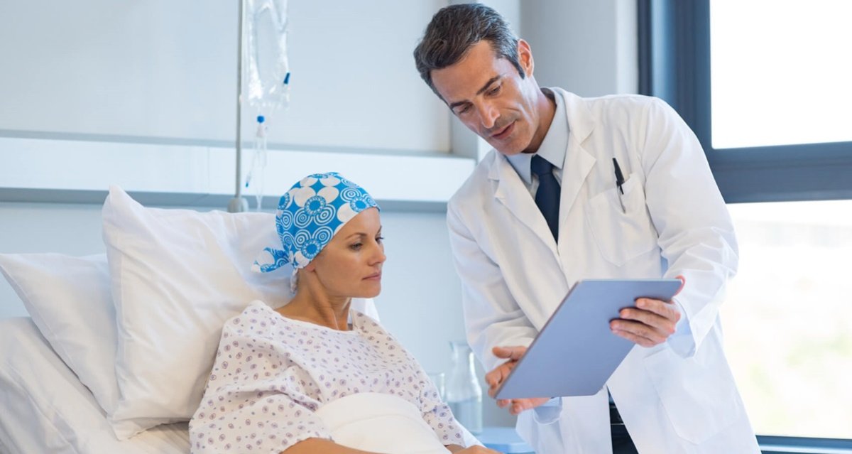 Eine Patientin mit Kopfbedeckung liegt in einem Krankenhausbett, schaut zusammen mit einem Arzt Unterlagen an, welche für die Chemotherapie relevant sind.