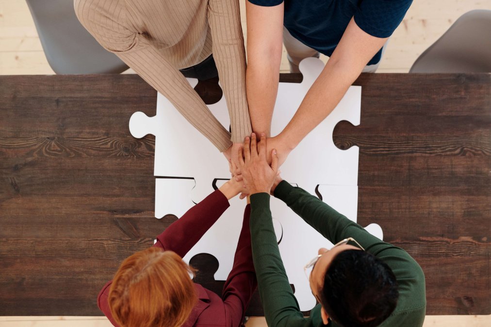 4 Personen halten über ein Puzzle die Hände zusammen. Motto: Hand in Hand arbeiten.