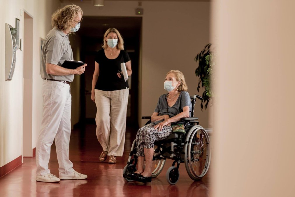 Betreuer steht vor einer Patientin im Rollstuhl und deren Angehörigen.