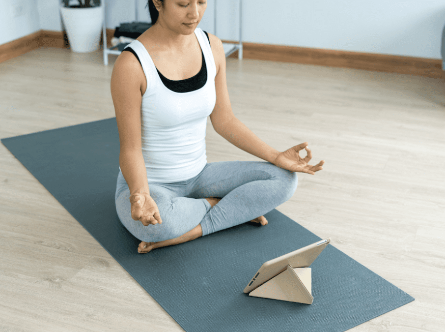 Junge Frau im Schneidersitz auf grauer Yogamatte vor einem Tablet.