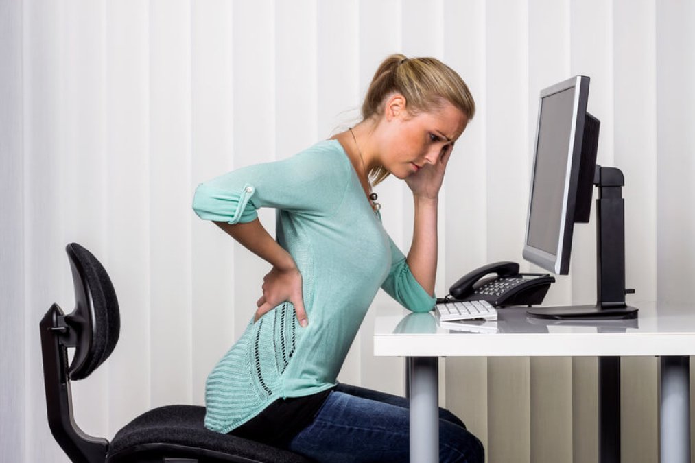 Frau hält sich den Rücken vor Schmerzen und sitzt am Schreibtisch vor einem Computer.