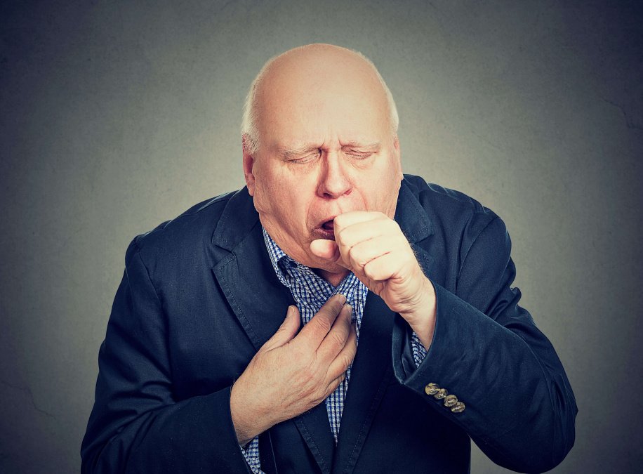 Ein älterer Mann im Anzug hält sich die Brust und eine Faust vor den Mund aufgrund eines Hustenanfalls durch COPD.