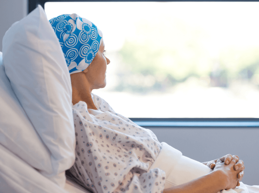 Krebserkrankte Frau mit blauem Kopftuch sitzt im Krankenbett und schaut aus dem Fenster.